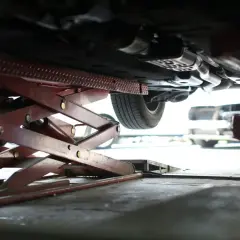 Podniesiony samochód na podnośniku nożycowym. Widać podwozie, wydech i koło. Mechanik sprawdza, co to jest tuleja w zawieszeniu.