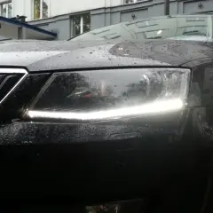 Czarne auto w deszczu, z włączonymi światłami dziennymi. Krople wody na masce i reflektorach tworzą ciekawy efekt.