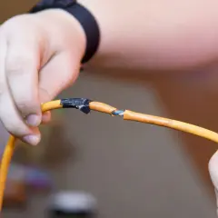 Dłonie trzymające pomarańczowy kabel, który jest przerwany. Widać niebieskie żyłki. Jak naprawić przerwany kabel? Taśma izolacyjna jest już nałożona.