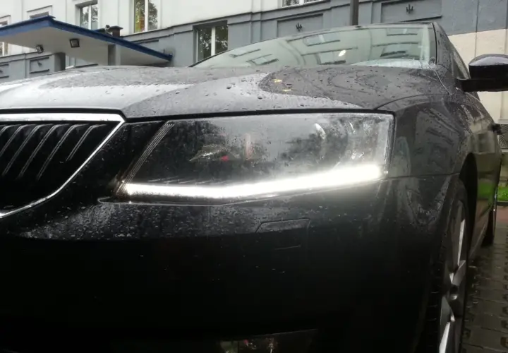 Czarne auto w deszczu, z włączonymi światłami dziennymi. Krople wody na masce i reflektorach tworzą ciekawy efekt.