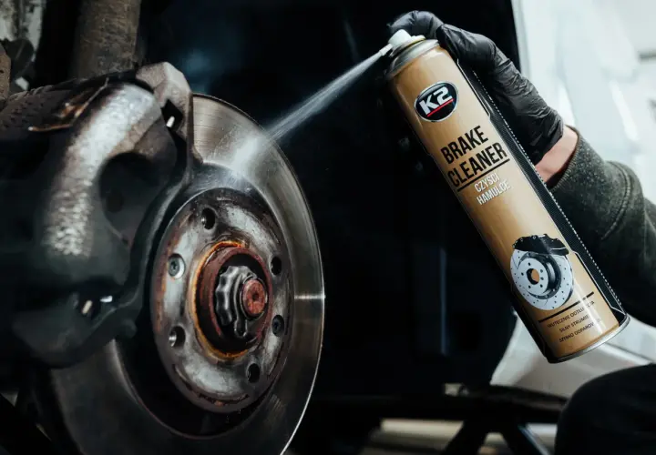 Rękawiczka w czarnej rękawicy rozpyla środek K2 Brake Cleaner na tarczę hamulcową, usuwając brud i rdzę.