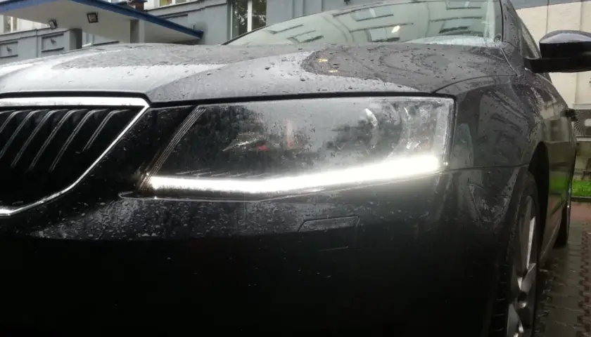 Czarne auto w deszczu, z włączonymi światłami dziennymi. Krople wody na masce i reflektorach tworzą ciekawy efekt.