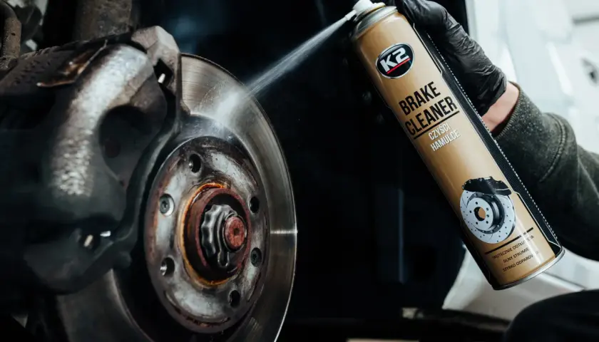 Rękawiczka w czarnej rękawicy rozpyla środek K2 Brake Cleaner na tarczę hamulcową, usuwając brud i rdzę.