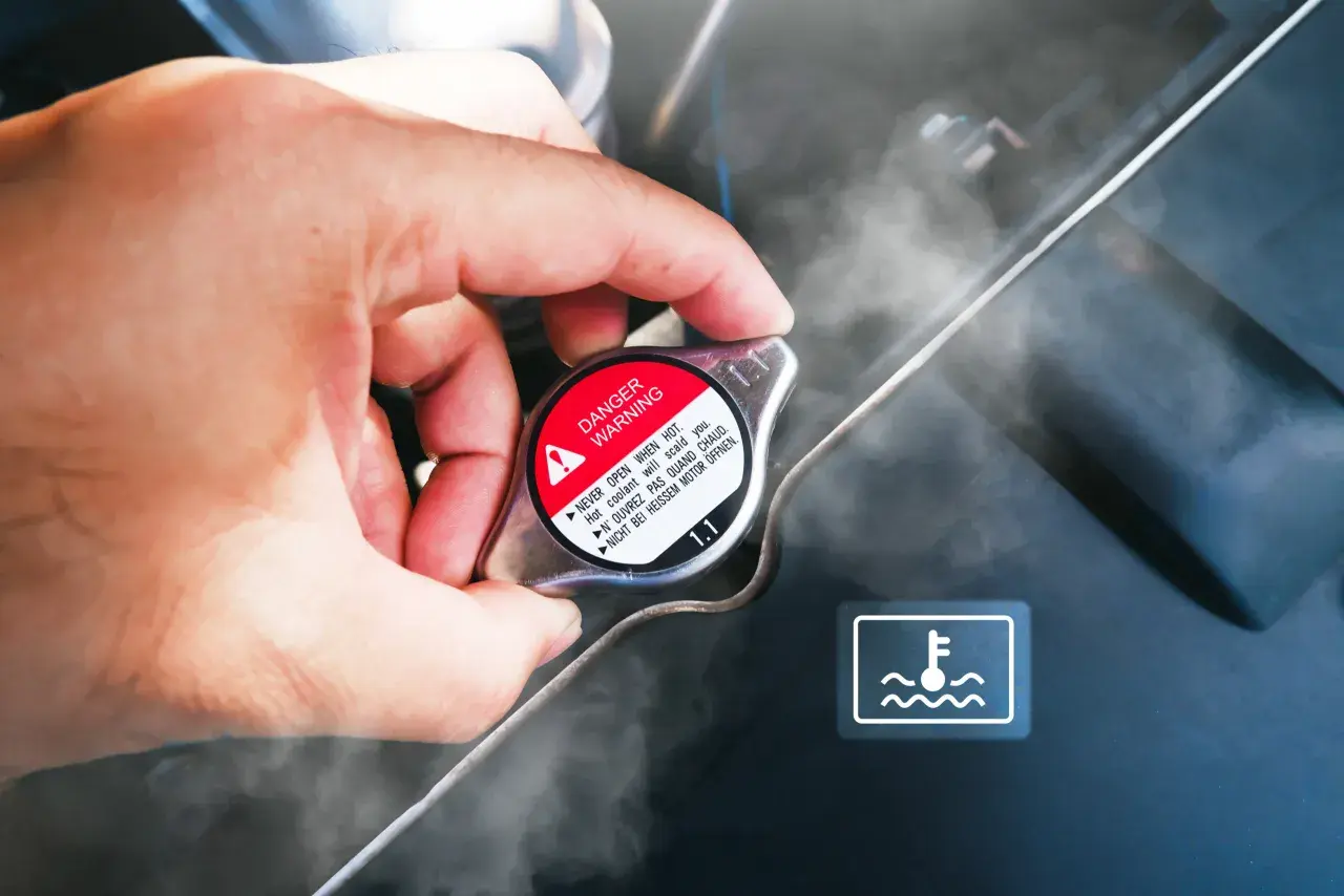Dłoń dotyka korka chłodnicy, z którego wydobywa się para. Widoczny symbol ostrzegawczy i napis "DANGER WARNING". Sygnalizuje to prawdopodobnie **mało płynu w chłodnicy**.