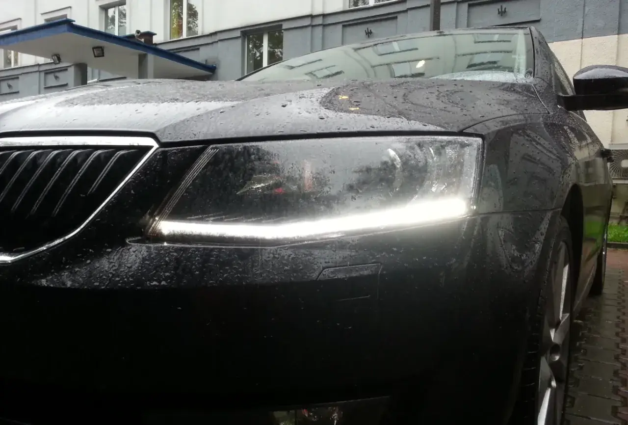 Czarne auto w deszczu, z włączonymi światłami dziennymi. Krople wody na masce i reflektorach tworzą ciekawy efekt.