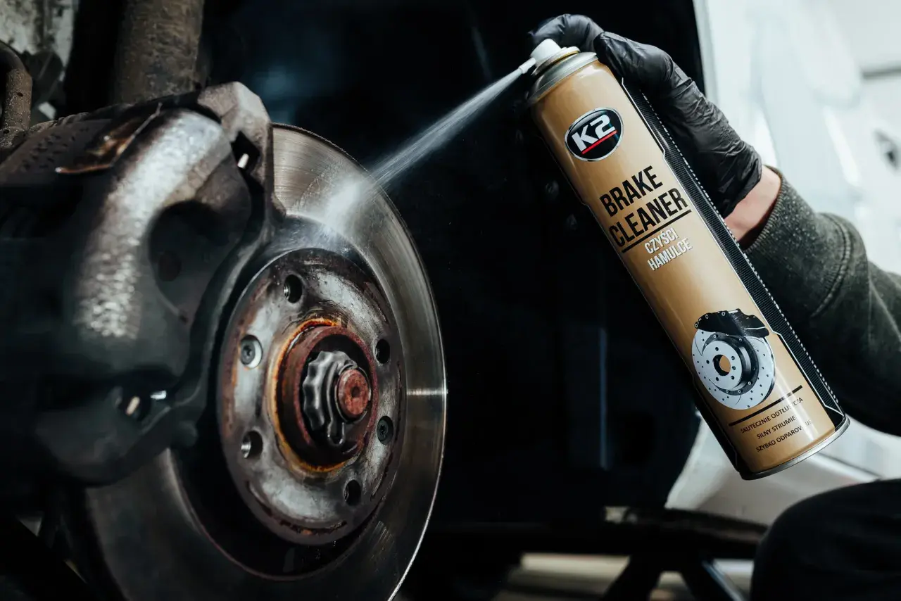 Rękawiczka w czarnej rękawicy rozpyla środek K2 Brake Cleaner na tarczę hamulcową, usuwając brud i rdzę.