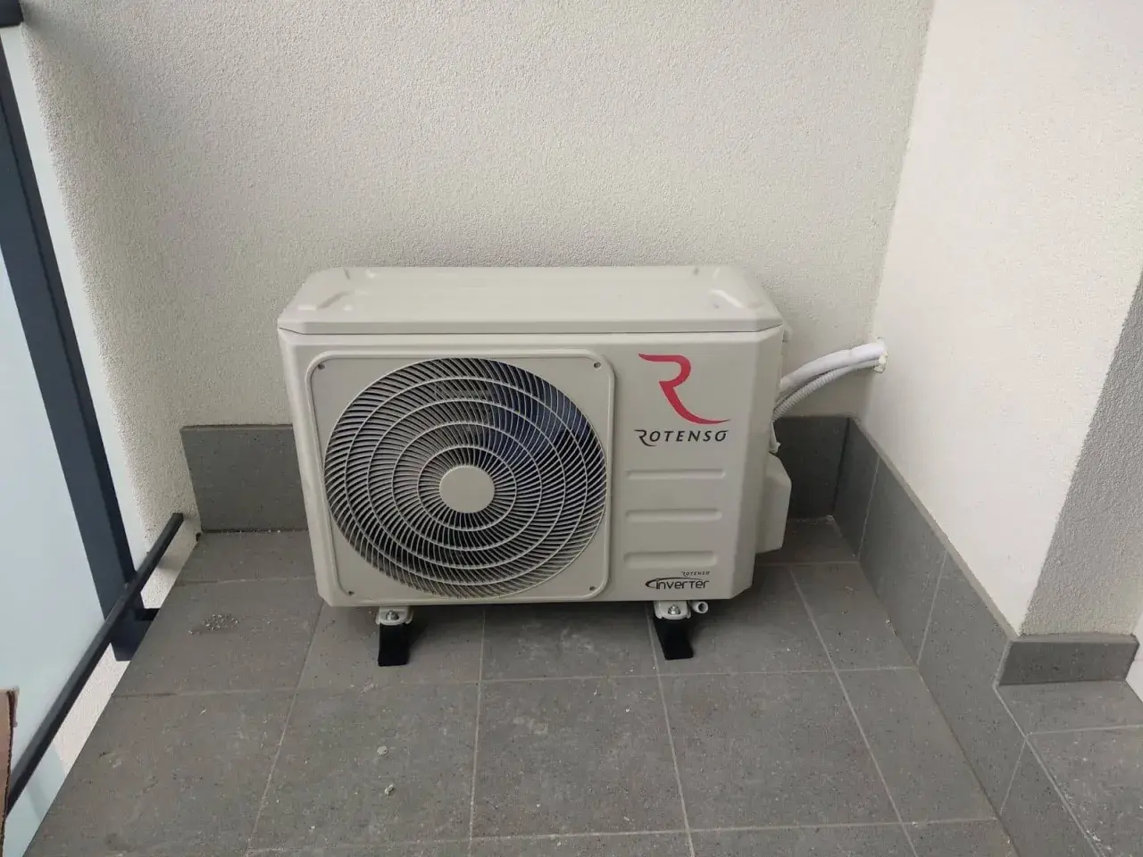 Jednostka zewnętrzna klimatyzacji Rotenso z logo, zamontowana na balkonie. Złącze klimatyzacji jest widoczne po prawej stronie.