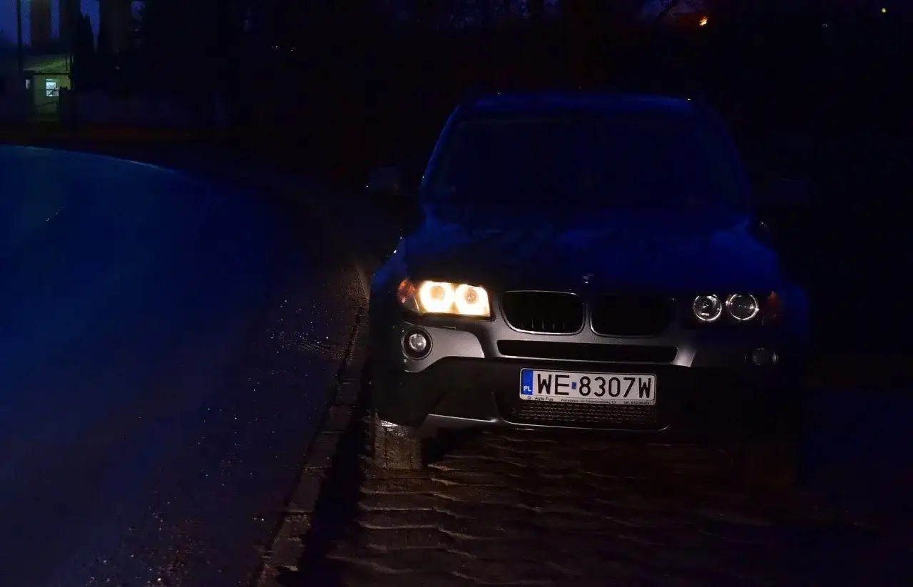 Ciemny SUV z włączonymi światłami pozycyjnymi i postojowymi stoi na mokrej, brukowanej drodze nocą.