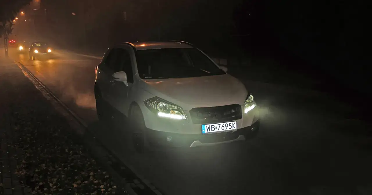 Biały SUV z włączonymi światłami dziennymi jedzie nocą przez mglistą drogę. Widać inne samochody z przodu.