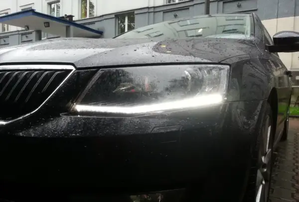 Czarne auto w deszczu, z włączonymi światłami dziennymi. Krople wody na masce i reflektorach tworzą ciekawy efekt.