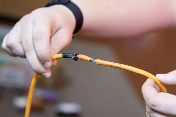Dłonie trzymające pomarańczowy kabel, który jest przerwany. Widać niebieskie żyłki. Jak naprawić przerwany kabel? Taśma izolacyjna jest już nałożona.
